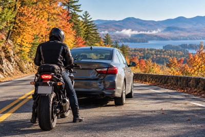 A motorcyclist stands safely beside a stopped motorcycle after a rear-end collision on a New Hampshire road.
